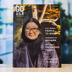 A copy of “Graphic Design USA” magazine depicting a woman wearing glasses and a jacket stands atop a white table.