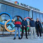 Aca Anderson, Johnathan Richardson, Jackson Faulknor, and Josh Minker stand in a row in front of the NBC Sports building with the colorful Olympic rings and a large sign displaying “NBC Sports.”