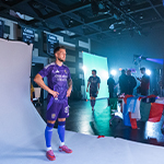 An Orlando City player poses for the camera with their hands on their hips wearing a purple uniform. Production crew members stand around in the background.
