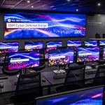 A room with dozens of desktop computers and chairs at long tables. A large screen on the wall reads Full Sail University IBM Cyber Defense Range Powered by AWS and Cloud Range.