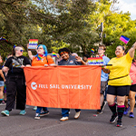 Full Sail students holding a Full Sail University banner march during the Pride Parade. A crowd holding rainbow signs and flags walks behind them.