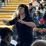 Students perform in a brass ensemble as a conductor leads, while audience members are seated in an amphitheater with Full Sail University tents in the background.