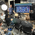 Shawn Rinehart stands in front of an audience of students and faculty next to a screen displaying numerous headshots. Lighting and camera equipment are around the room.