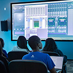 Students sit in a classroom watching a large screen displaying audio mixing software, with laptops open and studio speakers mounted on the walls.