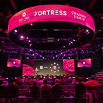 An audience sits in rows of chairs in a large room with a screen and stage. A speaker stands on the stage holding a microphone and red and purple lights shine.