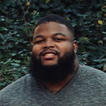 Tariq Ricks poses for the camera in front of a green leaf background. He is wearing a long sleeve grey shirt.