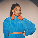DOE stands against a white wall. She has long braids tied up in a ponytail and wears a bright blue sweater and earrings with orange tassels.