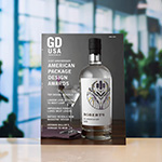 A copy of 'Graphic Design USA' magazine sits atop a white surface against a blurred background. The magazine cover depicts a bottle of gin and a martini glass against a gray background and reads '61st Anniversary American Package Design Awards.