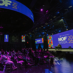 A speakers sits in a chair on a stage with a large screen behind them displaying “HOF Week 2026” and colorful graphics. An audience sits in rows of chairs facing the stage.
