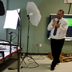 An instructor gestures while teaching in a studio setup with lights, cameras, and a green screen projection in the background.