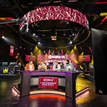 Analysts sit at a broadcast desk beneath a glowing “Madden NFL 26” ring as cameras roll on a live esports studio set.