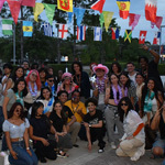 A diverse group of young adults poses outdoors under international flags, dressed festively with leis and fun accessories.