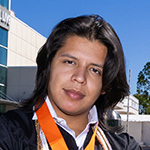 John Tobar stands with his arms crossed in front of the Full Sail Studios archway. He is wearing a graduation gown and an Armada medallion.