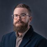 Drew Bailey wears a blue blazer and a brown checked shirt. He has brown hair, a beard, and glasses, and he’s smiling at the camera.