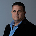 Sandy Navarro smiles at the camera in front of a grey background. He is wearing a blue button-down shirt and a black blazer.