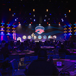 A crowd of people sit at round tables in a large room. A speaker stands on a stage holding a microphone with a screen behind them displaying the Full Sail University Veteran logo and logos of the branches of the military.