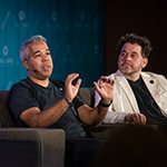 Juan Peralta and Gary Rizzo sit in armchairs on a stage and give a presentation. Juan wears a black t-shirt and Gary wears a black shirt and white blazer.