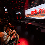 Students pack the rows of a live venue, watching a large screen presentation for ‘Drifter,’ with red stage lighting and a panel setup on stage.