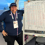 Alberto Pisani stands next to a large whiteboard with flight numbers on it and smiles. He wears a headset and a baseball cap that says Paris 2024.