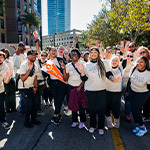 A large group of staff and students smile and wave at the camera wearing matching beige t-shirts. Buildings and trees are behind them.