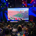 A crowd of students sit at round tables in a large room facing a large screen displaying a football stadium. One student wears a Puerto Rican flag as a cape.