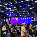 Students and alumni stand around and talk to potential employers. A screen behind them displays, “Welcome to the June 11th Career Fair.”