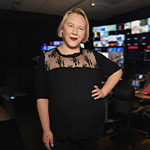 A person with blonde hair and a black lace-accent top stands in a dim control room, smiling, with colorful broadcast screens glowing behind them.
