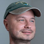 A portrait of a man wearing a green baseball cap and navy shirt while looking off to the side against a grey-green backdrop.