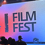 An audience faces a stage with a speaker holding a microphone. A large screen displays “Film Fest” and the VSU logo on a colorful background.