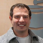 Nahun Paguada stands in front of a wall with a large sign displaying “The Latin Recording Academy” with a gramophone logo. He is wearing a white shirt, grey jacket, and khaki pants with his hands in his pockets.