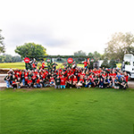 A large group of staff, faculty, and their families pose for a photo wearing red and green holiday shirts in front of a semi-truck bed decorated with garlands and lights. Some stand on the truck bed holding various musical instruments.