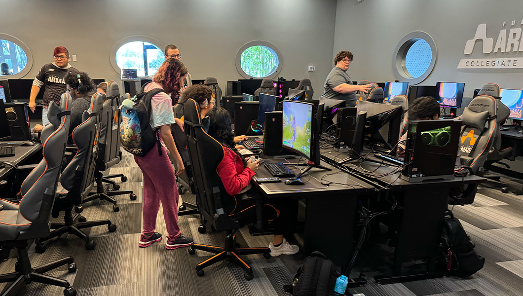 Several students sit at video game consoles in a large room as more students stand around and watch.