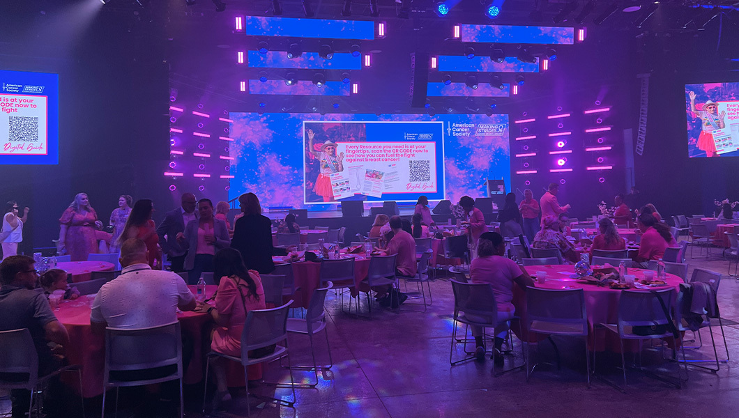 People sit at several round tables in a large room with a stage. Three screens display the American Cancer Society logo and pink and blue lights shine across the room.