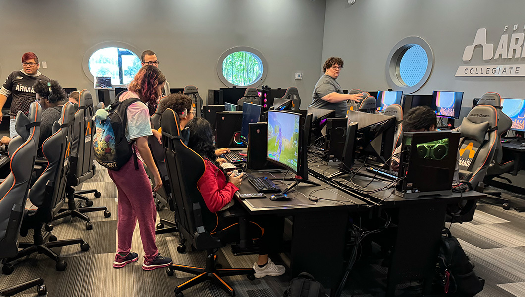 Several students sit at gaming consoles and play games while other students stand behind them and watch.