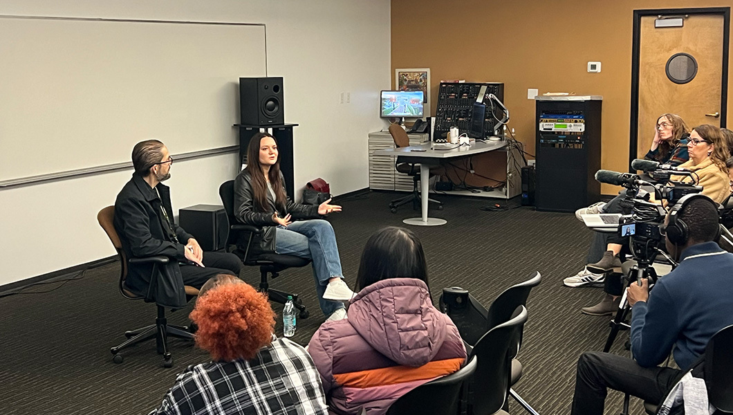 Beka Bishop sits in a chair and speaks to an audience of students in a classroom. Israel Vasquetelle sits next to Beka and moderates the talk.
