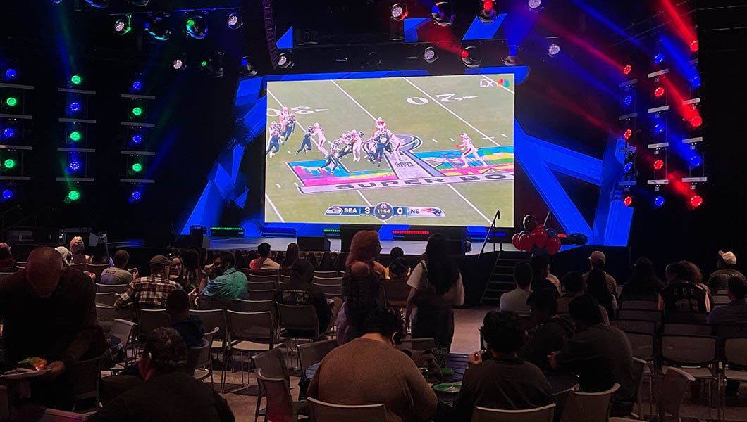Students sit in rows of chairs facing a large screen displaying a football field and players playing football.