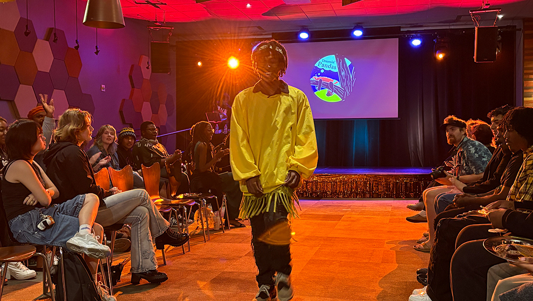 A student smiles as they walk the runway wearing a yellow sweatshirt and black jeans. The audience sits in chairs on either side of the runway facing the model and clapping and cheering.