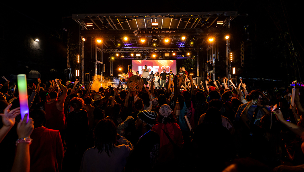 The Outdoor Stage is lit up with lights and a screen displaying “HOF Week 2026” as students stand together in a crowd with their hands raised and holding glow sticks.