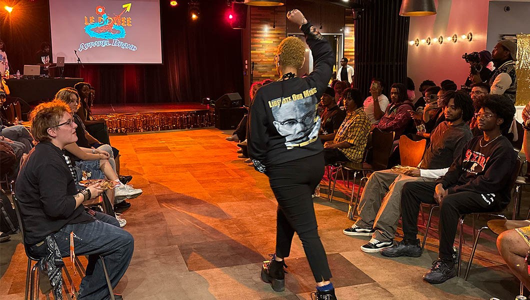 A student walks the runway wearing black boots, pants, and a sweatshirt with their fist raised in the air as the audience watches.
