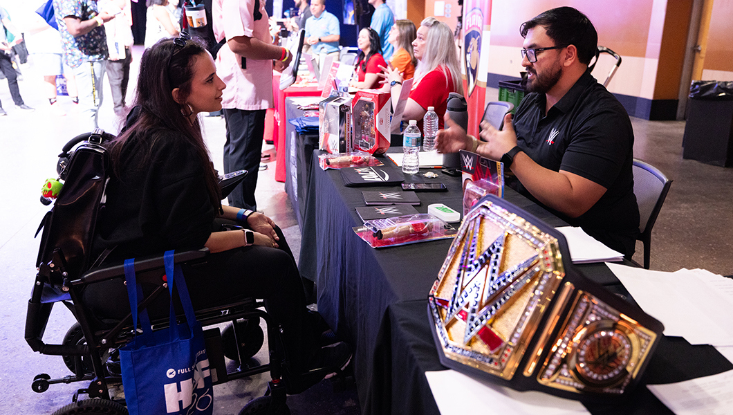A student in a wheelchair talks to a WWE representative across a table with a gold WWE belt next to them. Other people talk in the room around them.