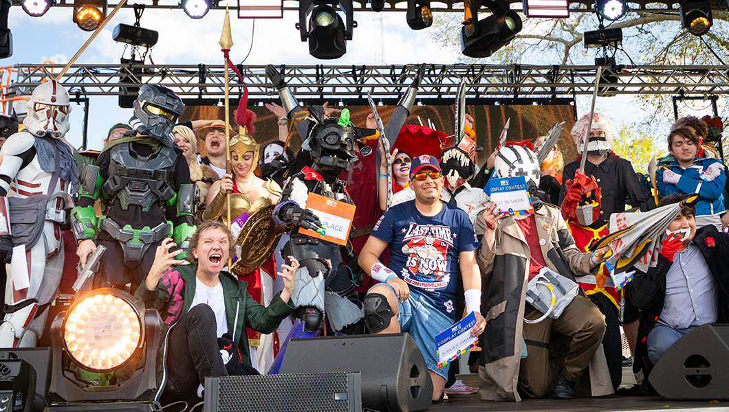 A large group of students in costumes pose for a group photo on the Outdoor Stage. They’re costumes include Stormtroopers, John Cena, La Muerte from The Book of Life, and many more.