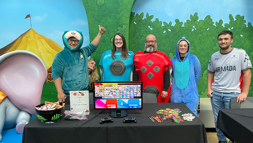 Staff and students in costumes stand behind a table with a bowl of candy, stickers, and other goodies on it, as well as a gaming console and two controllers.