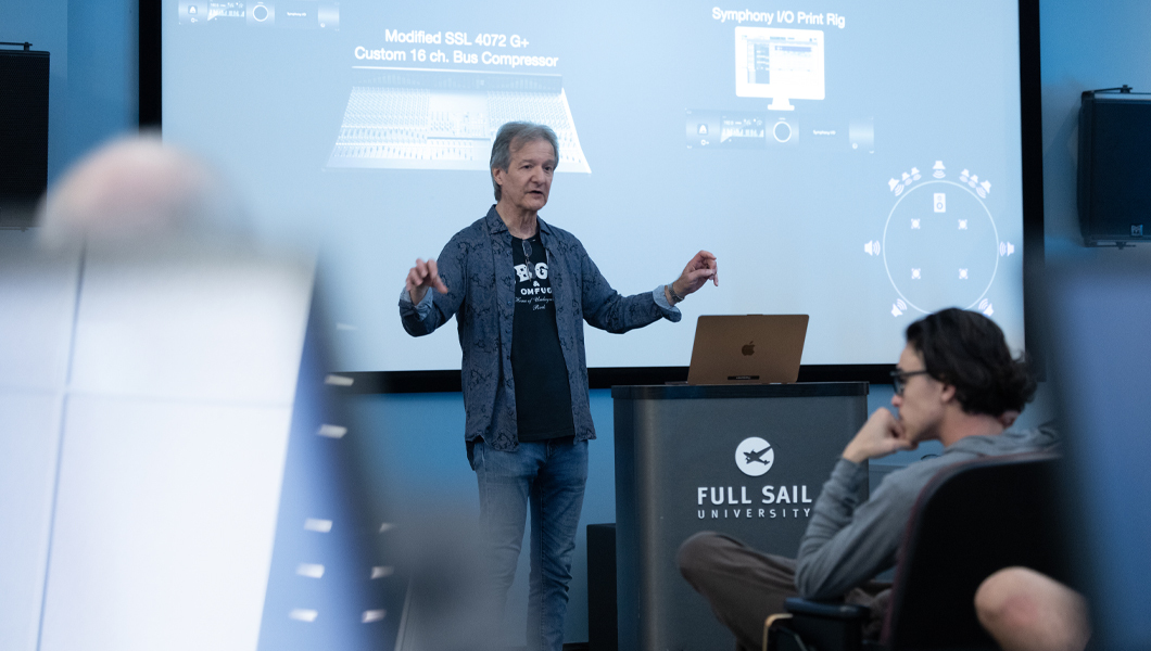 Bob Clearmountain speaking at Full Sail University in front of a presentation screen while standing next to a laptop on a podium, with an attentive audience in the foreground.
