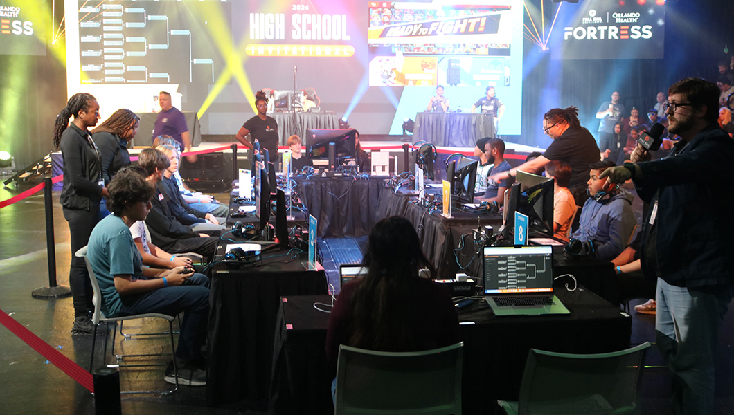 A gaming tournament is underway with players seated at monitors, a commentator speaking, and a lively audience in a brightly lit arena.