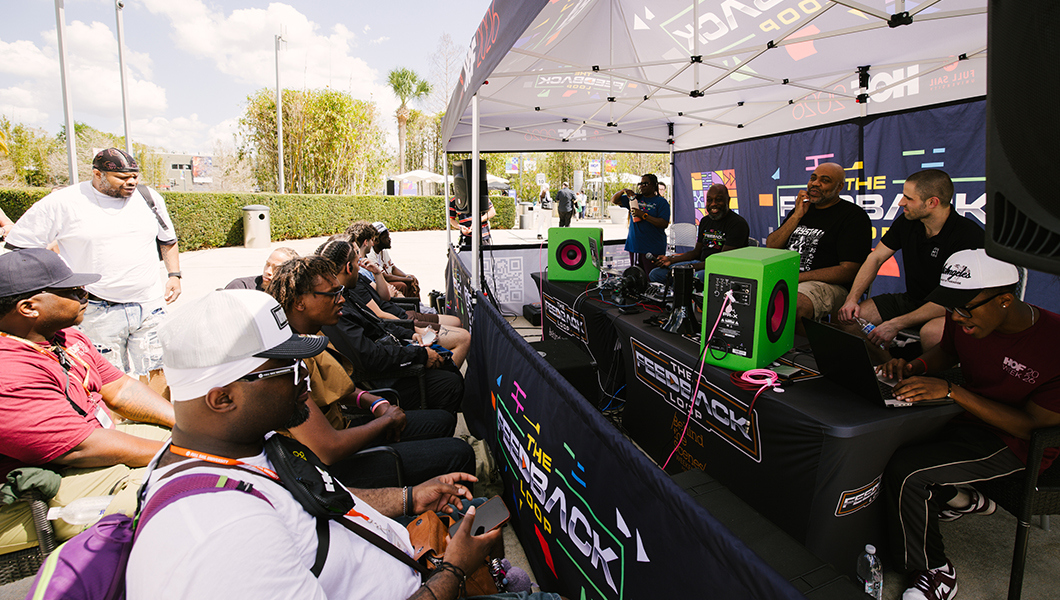 Students sit in chairs and face a tent where Leslie Brathwaite and Tremaine Williams sit at a table with microphones and bright green speakers. A sign at the front of the tent displays “The Feedback Loop” with colorful graphics.