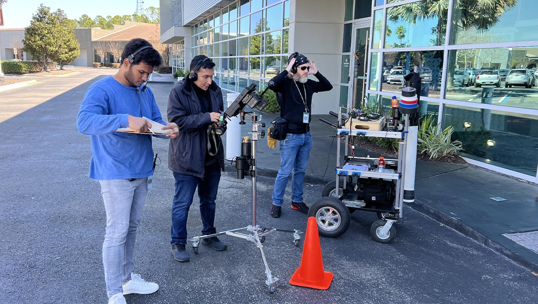 Two students and a faculty member wearing headsets stand outside of a building. One holds a clipboard, one holds a camera, and one adjusts their headset. A cart holding gear is next to them and an orange traffic cone is on the ground.