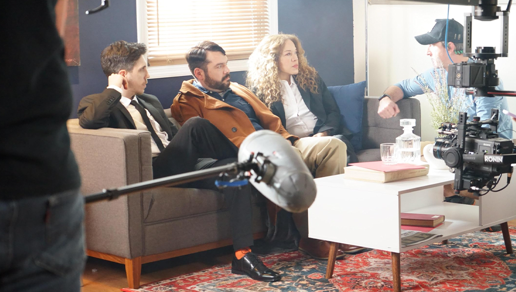 Three actors sit on a grey couch with a white coffee table across from them and a red rug on the floor. A crew member crouches next to the couch and speaks to the actors and a camera operator angles a camera at them.