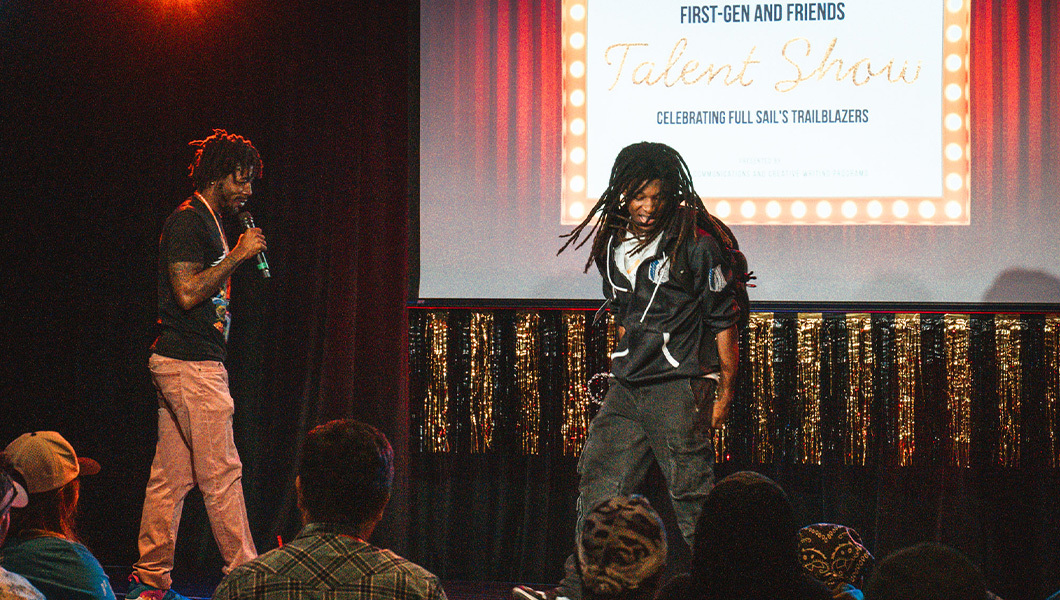Two students take the stage and perform in front of an audience. One speaks into a microphone and one dances. A screen behind them reads “First-Gen and Friends Talent Show Celebrating Full Sail’s Trailblazers.”