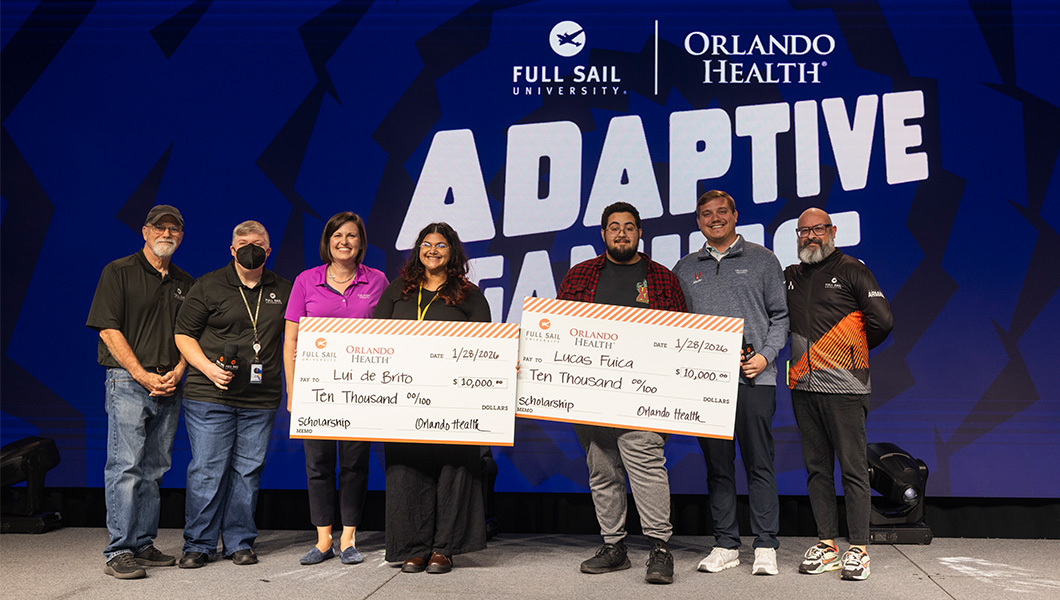 Staff members of Full Sail and Orlando Health stand onstage next to Lui de Brito and Lucas Fuica, who are holding giant checks for $10,000 each. Behind them a large screen displays “Adaptive Gaming” and the Full Sail University and Orlando Health logos.