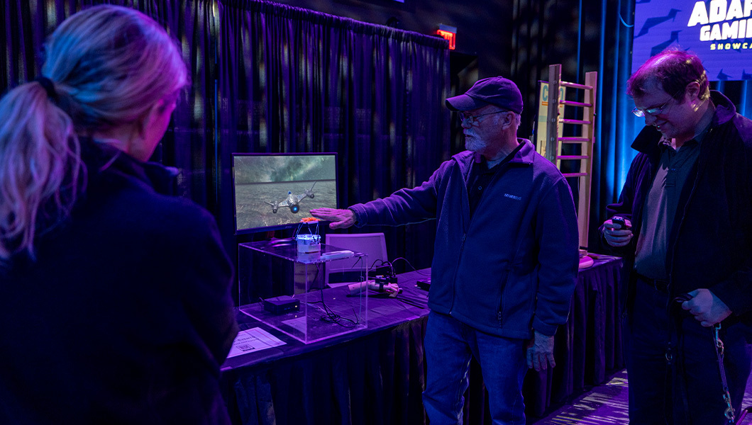 Rob Catto holds his hand over a motion controller connected to an airplane flying game displayed on a nearby screen. Two people stand nearby and watch.